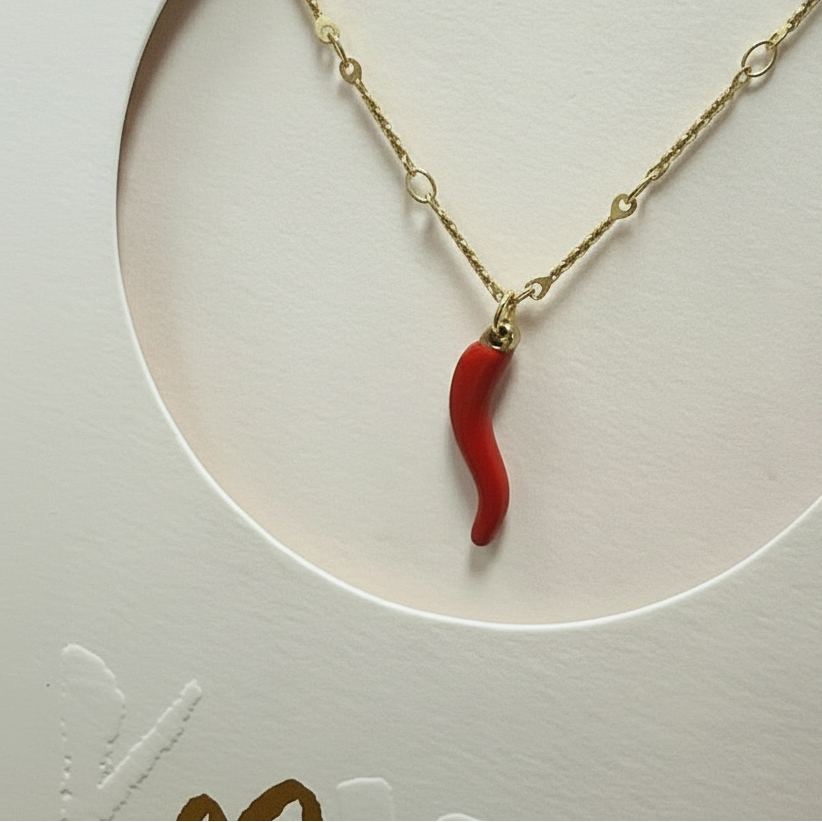A gold chain necklace with a small curved red chili pepper pendant, displayed inside a circular cutout on a textured cream-colored card.