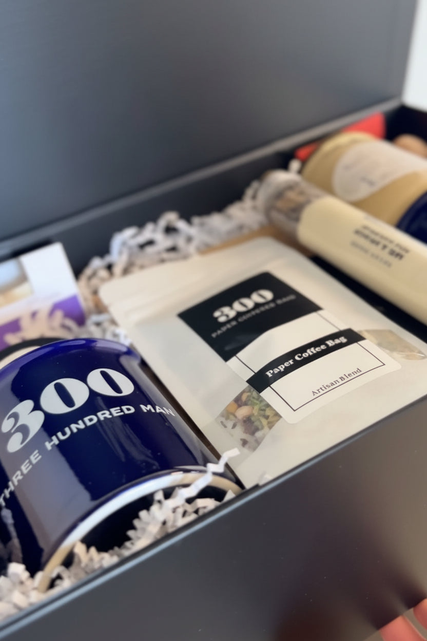 A dark gray gift box with 'Welcome Home' printed on the inside lid, containing a navy blue mug with 'Three HUNDRED MAIN' text, a white branded pouch, and other packaged items. A hand is visible holding the front of the box.