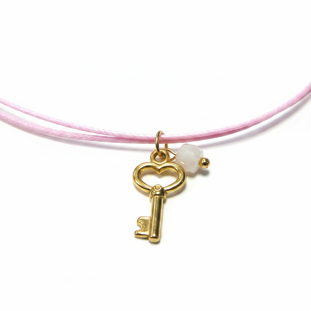 A gold key-shaped charm with a heart-shaped top and a small faceted white bead, attached to a light pink cord bracelet. The bracelet is laid on a white card with gold foil hearts and faint script text.