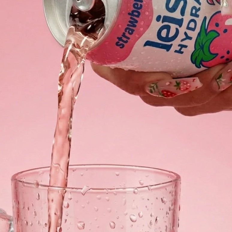 Leisure Hydration strawberry breeze can being poured into a glass with ice and strawberries on a pink background