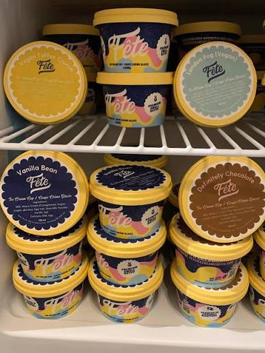 Multiple tubs of Fete ice cream arranged on white refrigerator shelves. Visible flavors include Vanilla Bean, Chocolate, and Vegan Chocolate, with yellow lids and dark blue labels.