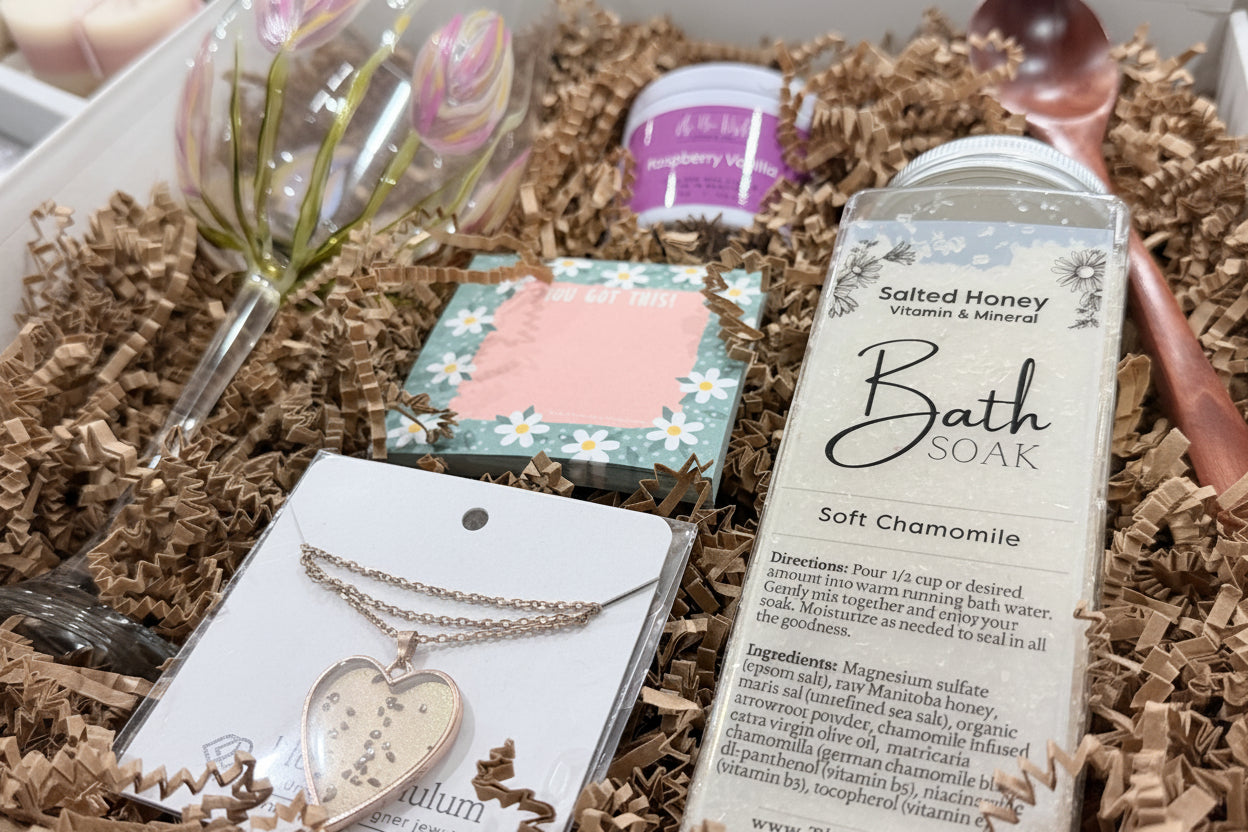 A bath gift set arranged in brown paper shred, featuring a large bottle of Salted Honey Bath Soak in Soft Chamomile, a pink and white floral soap bar, a small purple jar, a heart-shaped gold necklace on a card, and a clear glass ornament with pink tulips.