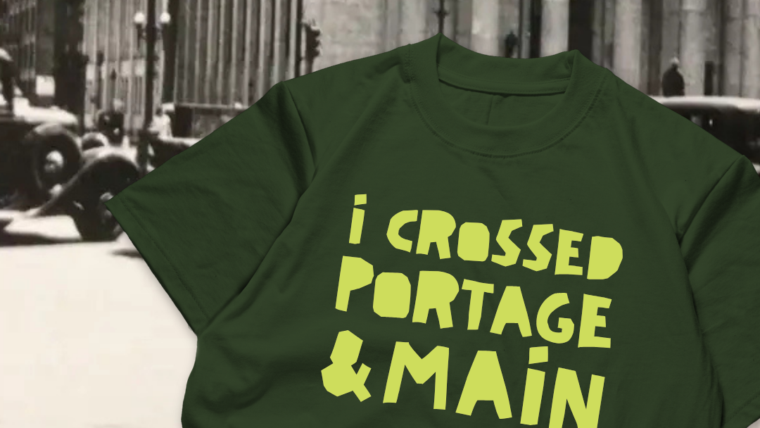 A dark green crewneck t-shirt with the phrase 'i CROSSED PORTAGE & MAIN' printed in a bold, yellow, retro-style font across the front, set against a blurred black and white street background.