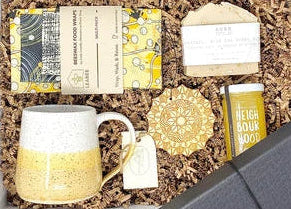 A curated gift box set displayed in an open black gift box filled with shredded brown paper. The set includes a speckled yellow and white ceramic mug, a packaged item with yellow patterned wrapping, a small white card, a gold-toned decorative piece, and a yellow cylindrical tin.