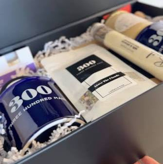A dark gray gift box with 'Welcome Home' printed on the inside lid, containing a navy blue mug with '300+ HUNDRED MAIN' text, a white branded pouch, and other packaged items. A hand is visible holding the front of the box.